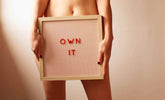 Own It framed sign held by a person, featuring red letters on a striped background.